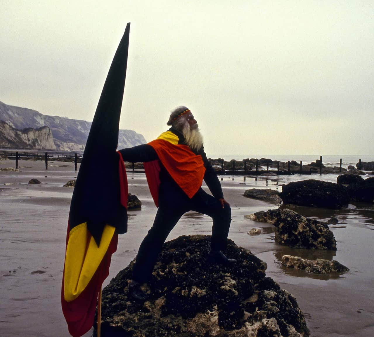 an aboriginal man holding an aboriginal flag stands on a rock at a dover beach, the white cliffs in the background, as he looks heroically out to sea