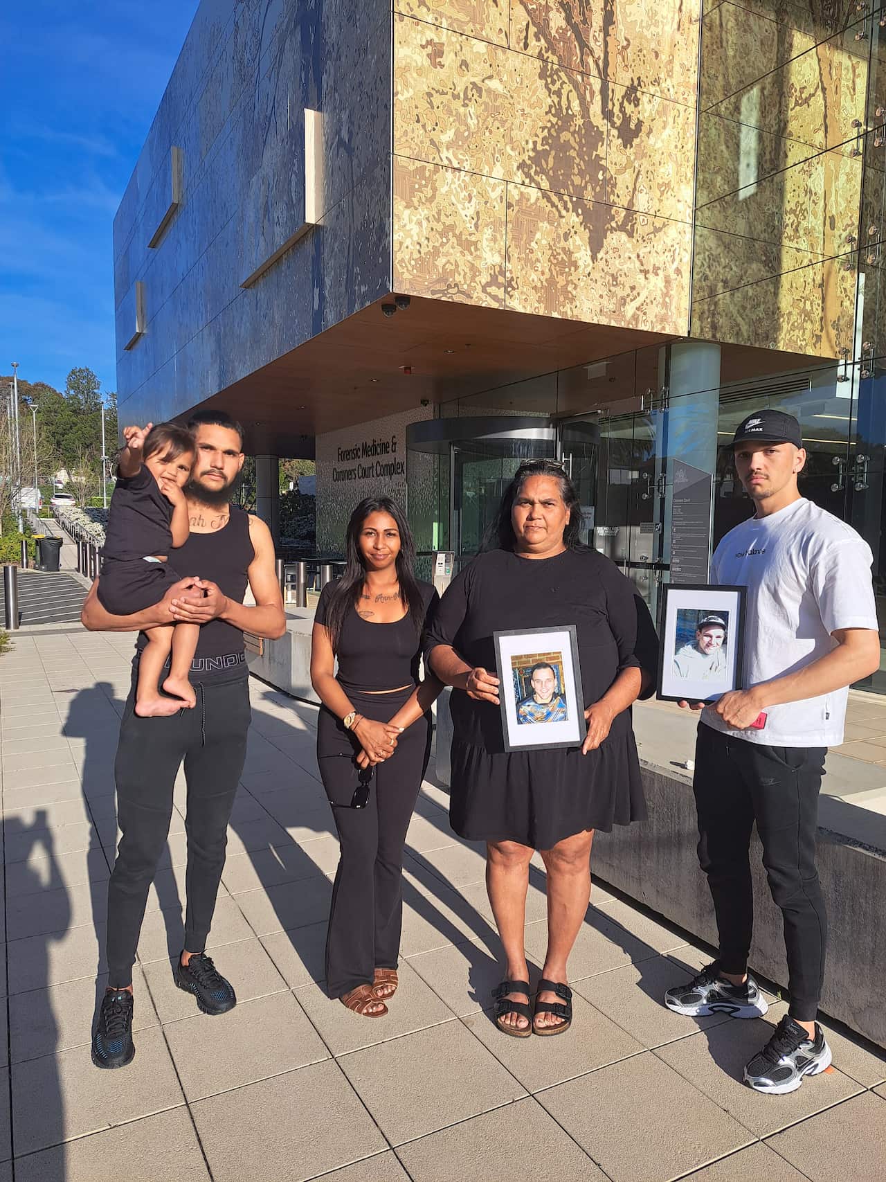Two men, two women and a child stand outside the Coroners Court holding two photos of their family member TJ.