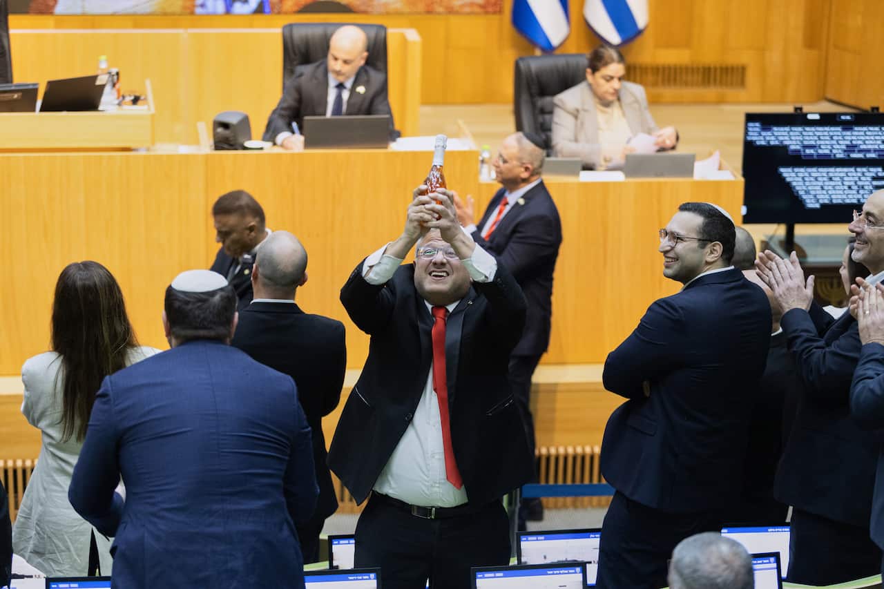 A man wearing a suit and a red tie holds his hands up as he celebrates inside a parliament chamber surrounded by other members