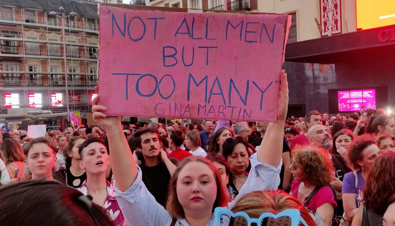 A woman standing in a crowd of people holds a sign above her head. It reads: Not all men but too many Gina Martin