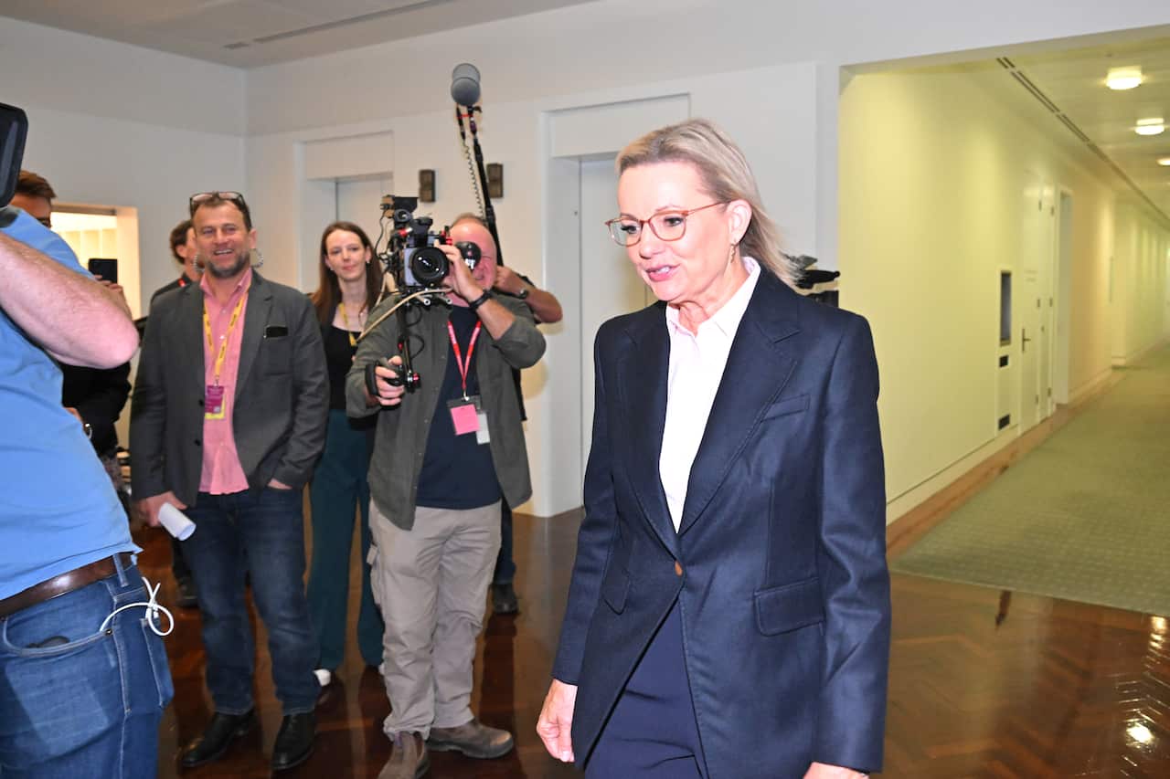 A woman in a blue suit is walking in a lobby with media persons following her.