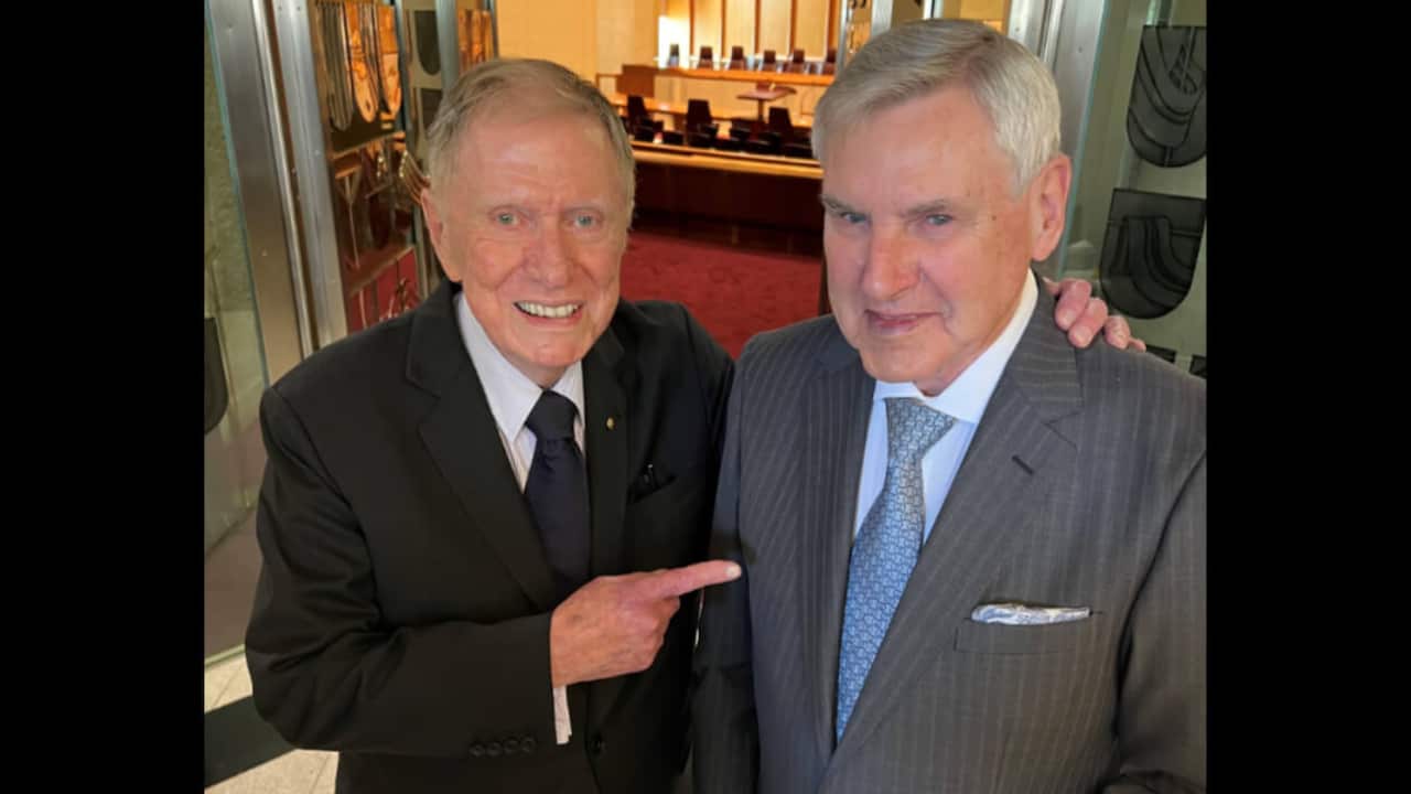 A man in a black suit with his arm around a man in a grey suit. Both are smiling
