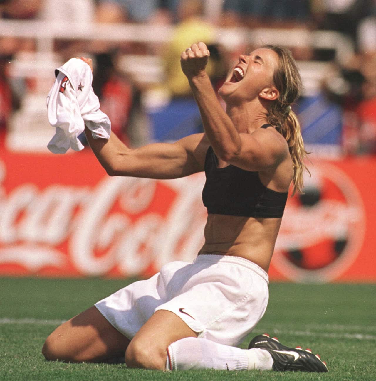 A woman in white shorts kneeling on the ground with arms out in celebration holding her shirt.