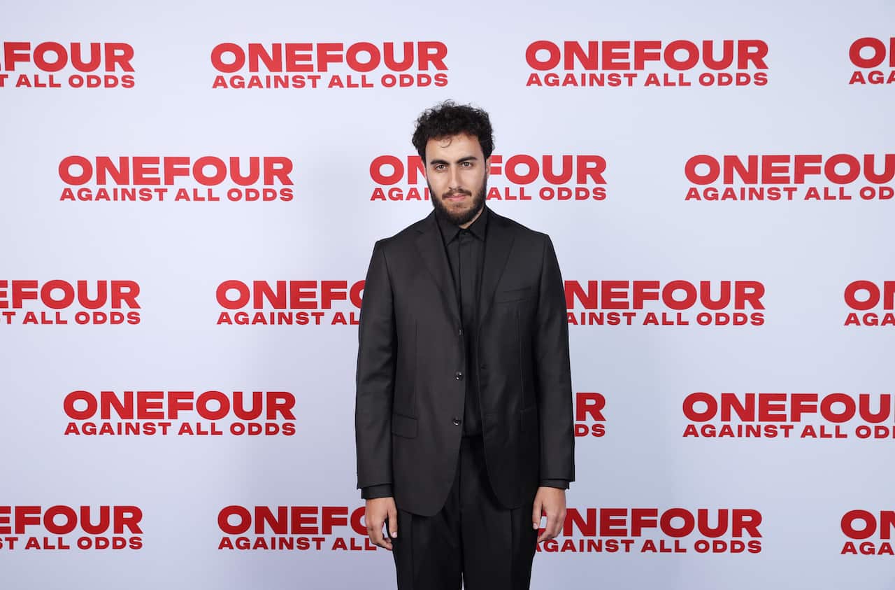 A man wearing a Black suit with a neutral expression is standing in front of a white screen with the words 'ONEFOUR Against All Odds' printed across the screen
