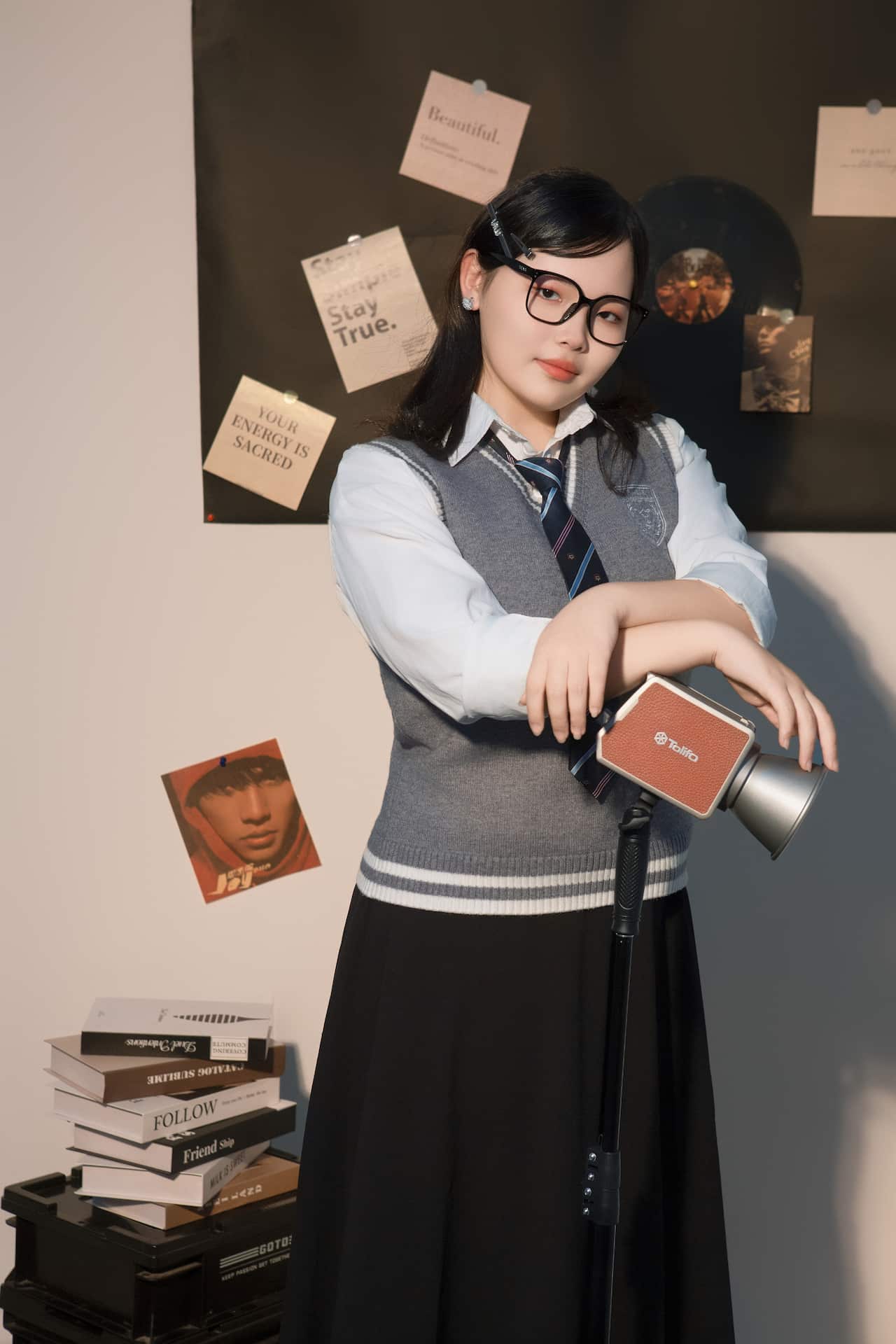 A young woman with glasses and a school uniform leans on a studio light in front of a dark backdrop decorated with notes and records.