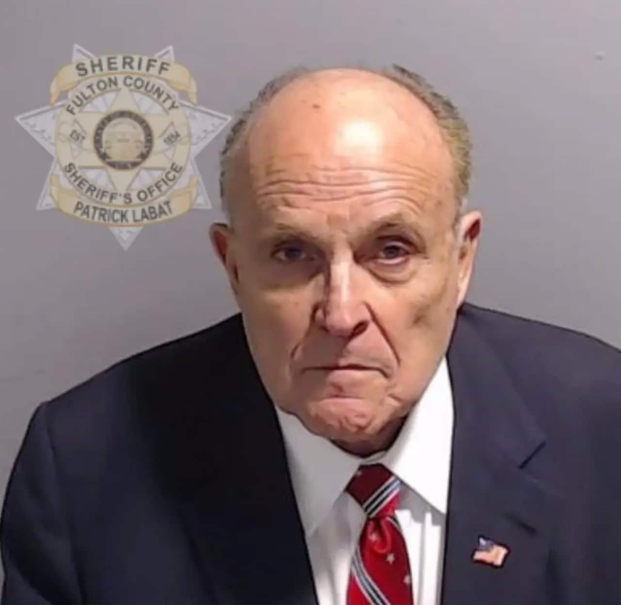 A man in a navy suit with an American flag pin on it posing for a mugshot.