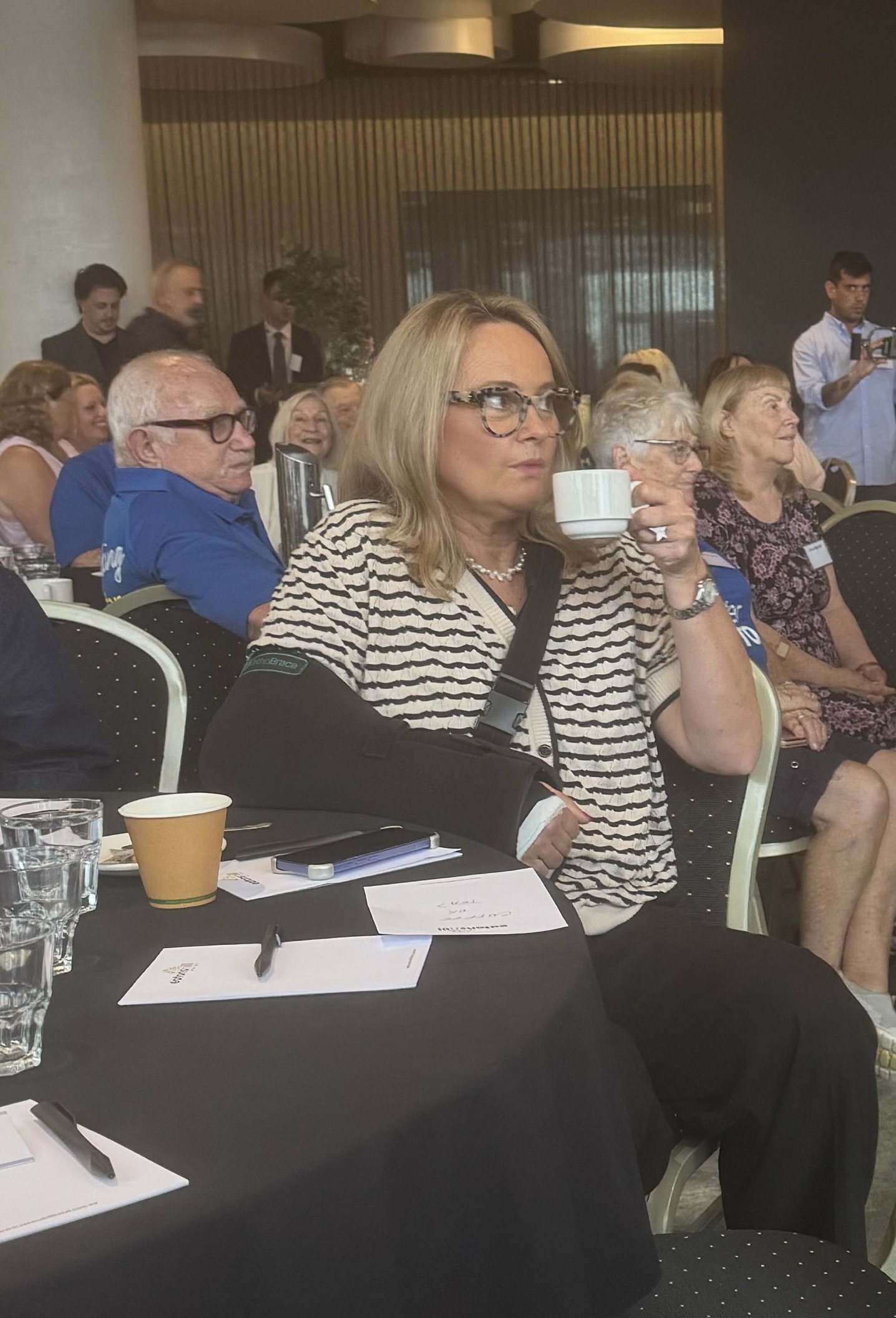A woman wearing a sling around her arm holds a cup in her other hand