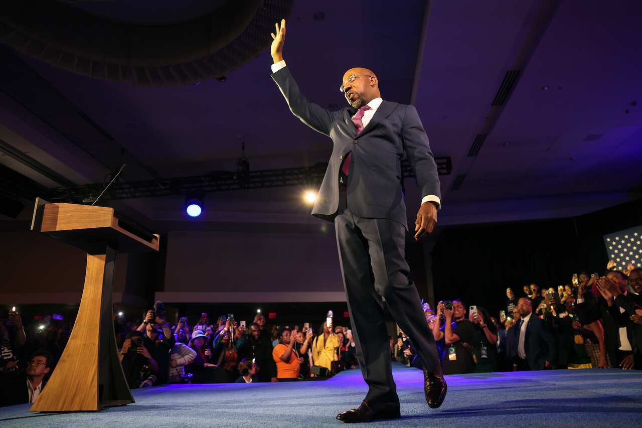 A man wearing a suit and tie waves as he walks toward a podium as a crowd cheers him on.