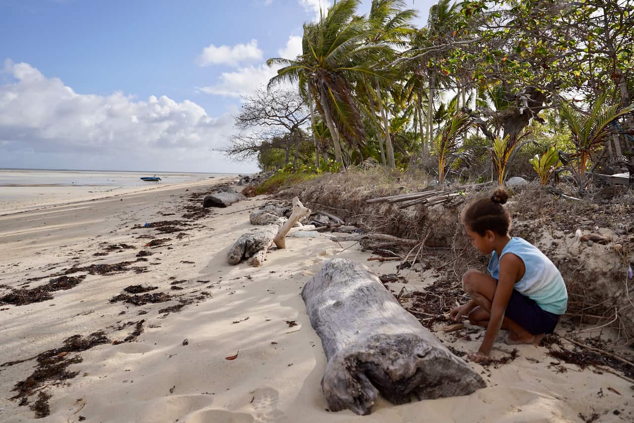 A young Kulkalgal boy wearing a pale blue singlet and dark shorts squats on a sandy beach.