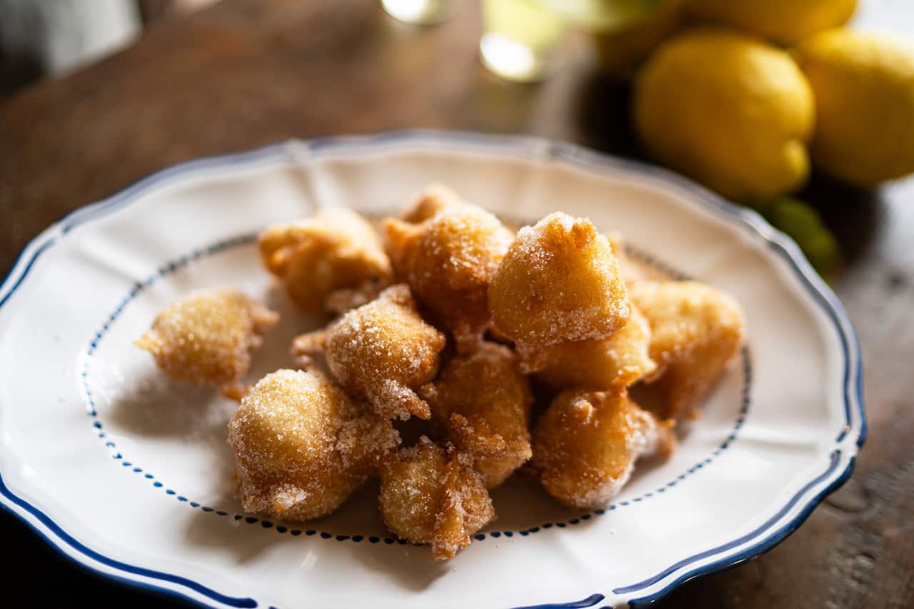 A pile of golden-brown fritters, dusted with sugar, sit on a blue and while plate.  