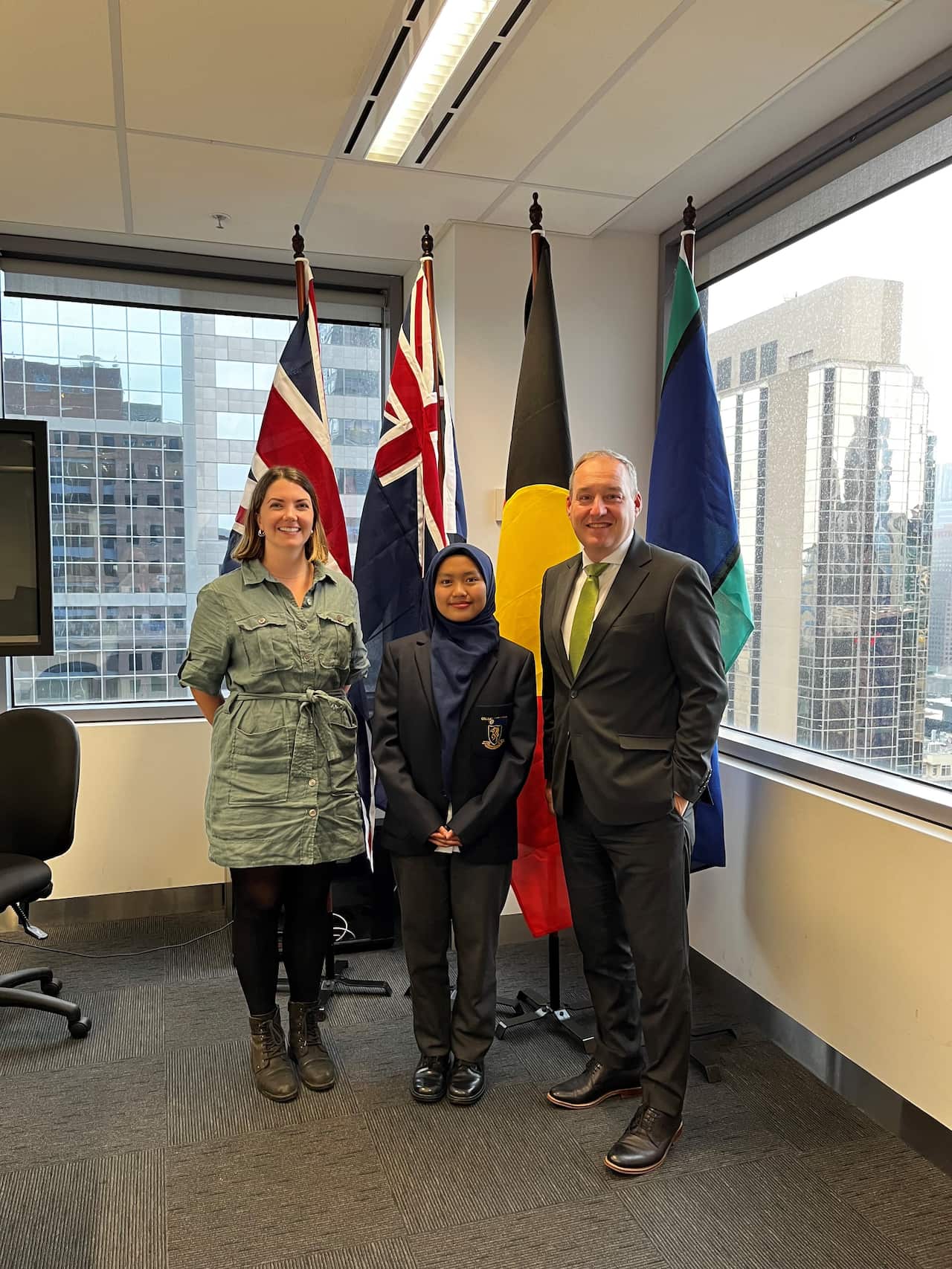 A young woman in a dark outfit stands between a man and a woman in front of the British, Australian, Aboriginal and Torres Strait Islander flags
