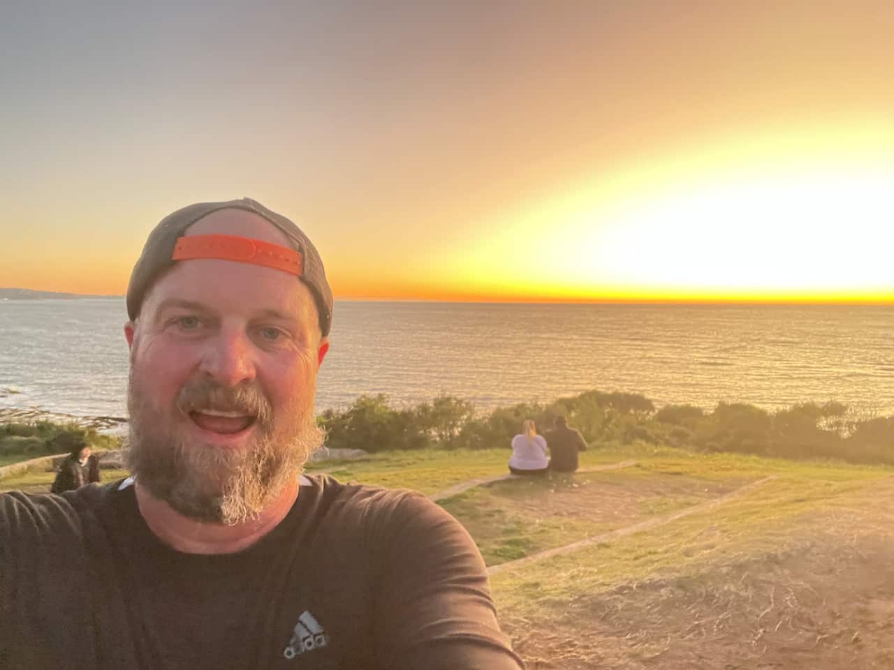 A smiling man takes a selfie. He is wearing a baseball cap backwards and there's a grassy cliff and the sea in the background.