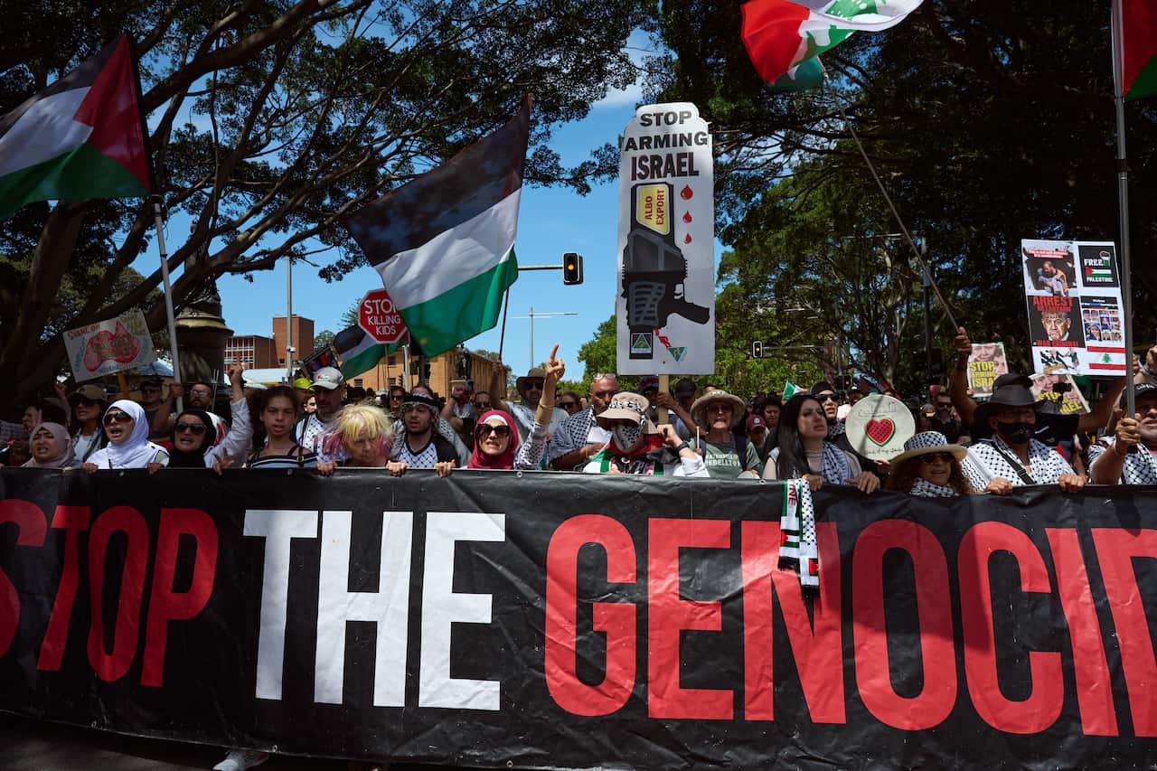 A large group of protesters outside walking behind large a banner that reads Stop the Genocide. 