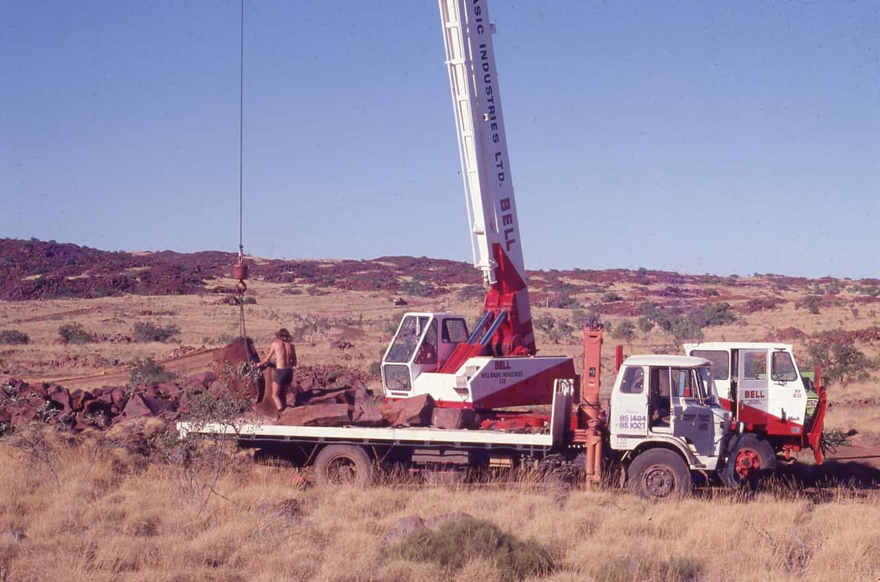 A mobile crane truck parked in an arid, rocky landscape while a worker maneuvers large rocks attached to a crane hoist.