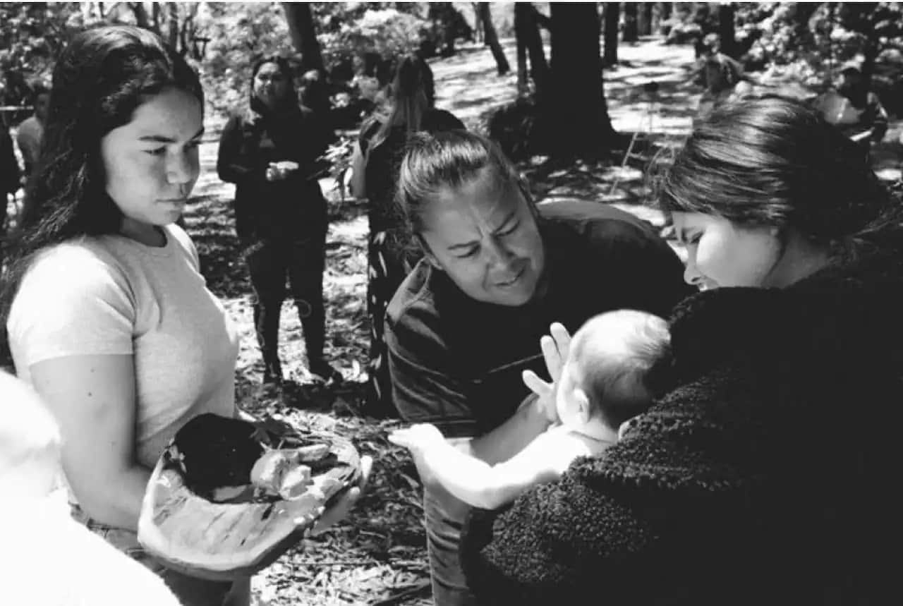 black and white photo of three Indigenous women standing in the bush, one holding a coolamon, another holding a baby, and the third applying traditional paint to the child. 