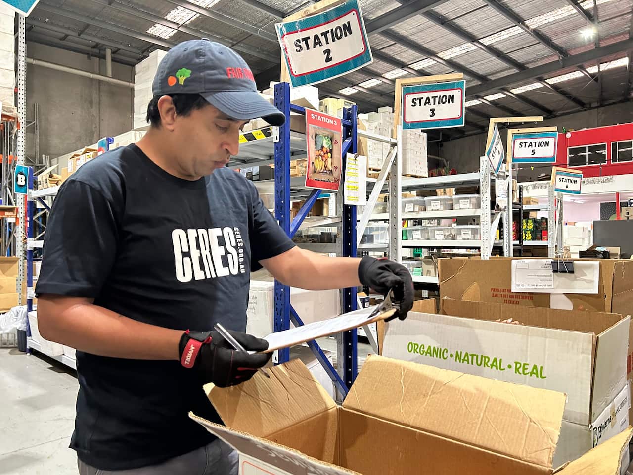 A man in a black t-shirt holding a clip board stands in a warehouse. 