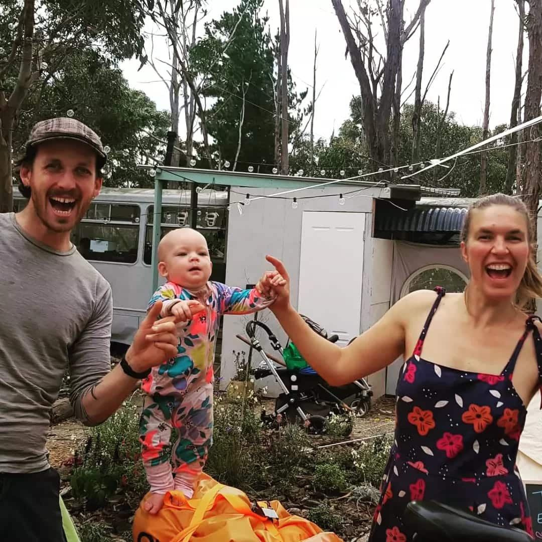 A man in a flat cap and a woman in a floral dress smile widely while an infant balances on a backpack between them, holding both their hands