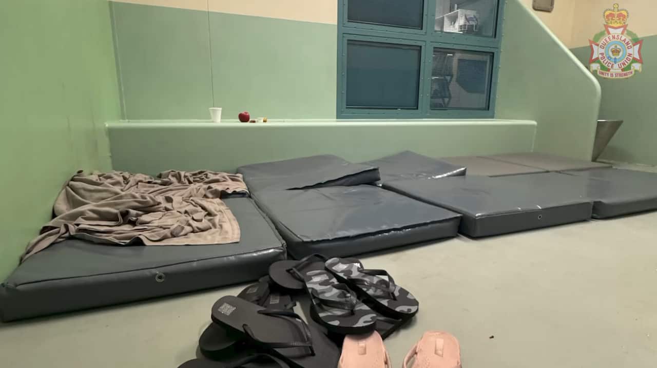 A pile of children's-sized thongs in front of ten thin, grey mats laid out on a concrete floor. The room has pale green walls, food scraps and a single blanket.