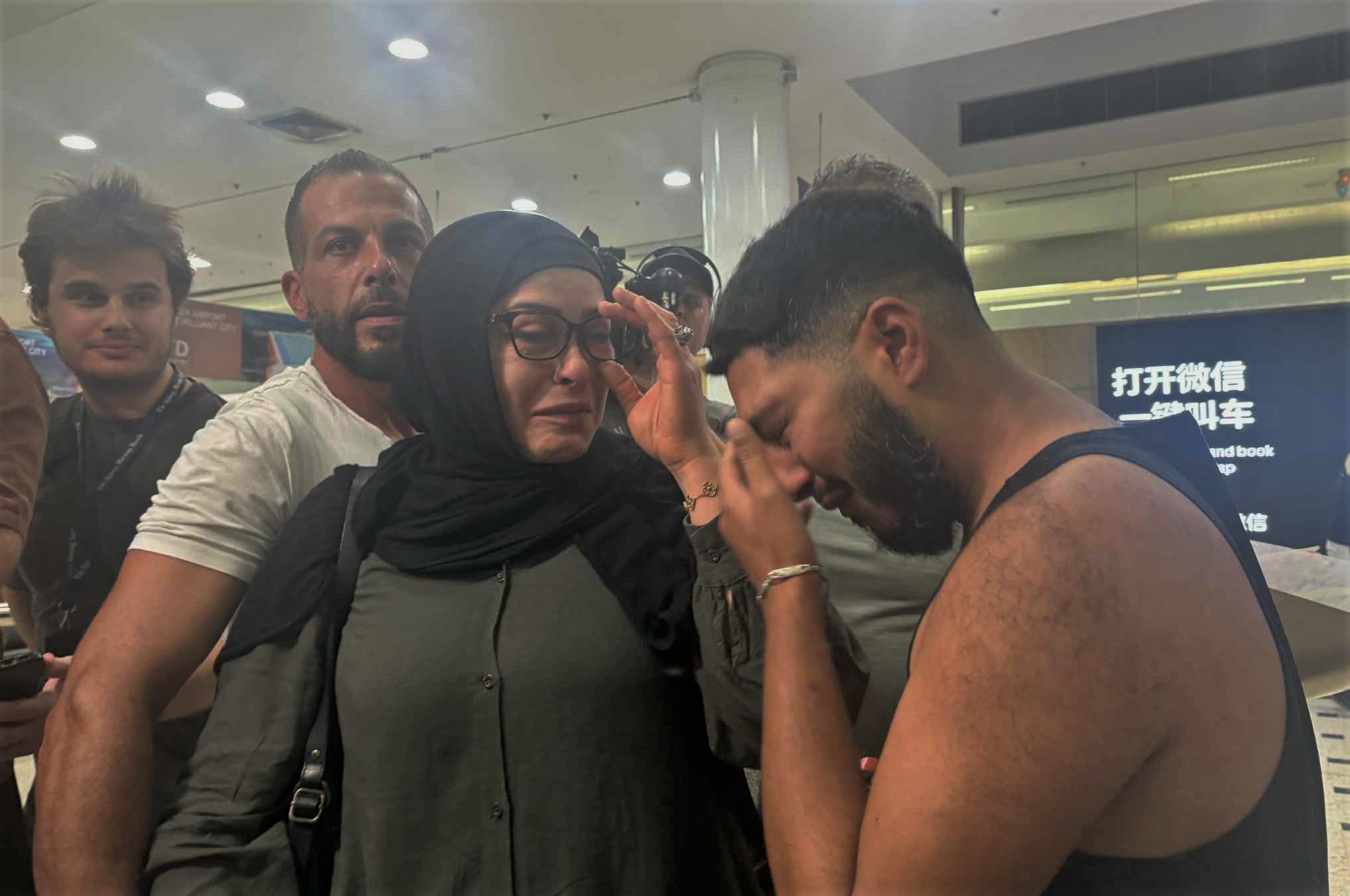 A woman wearing olive green with a black hijab and glasses appearing emotional next to a young man wearing a singlet crying into his hand.