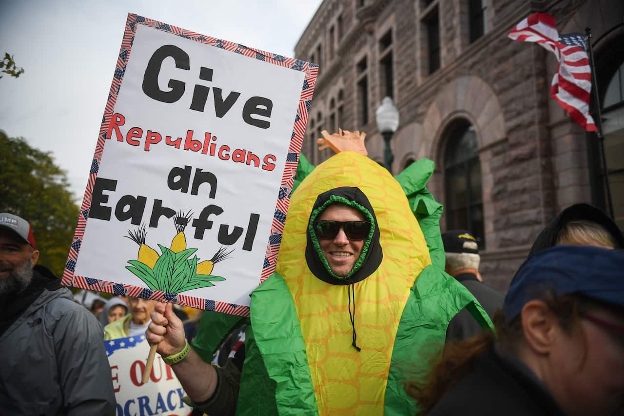 A person wearing an inflatable corn-on-the-cob costume holds a sign that reads, "Give Republicans an Earful," with an image of corn at the bottom.