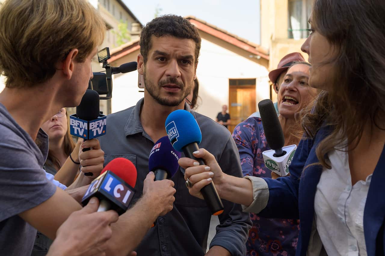 A serious looking man is surrounded by media, some holding microphones. 