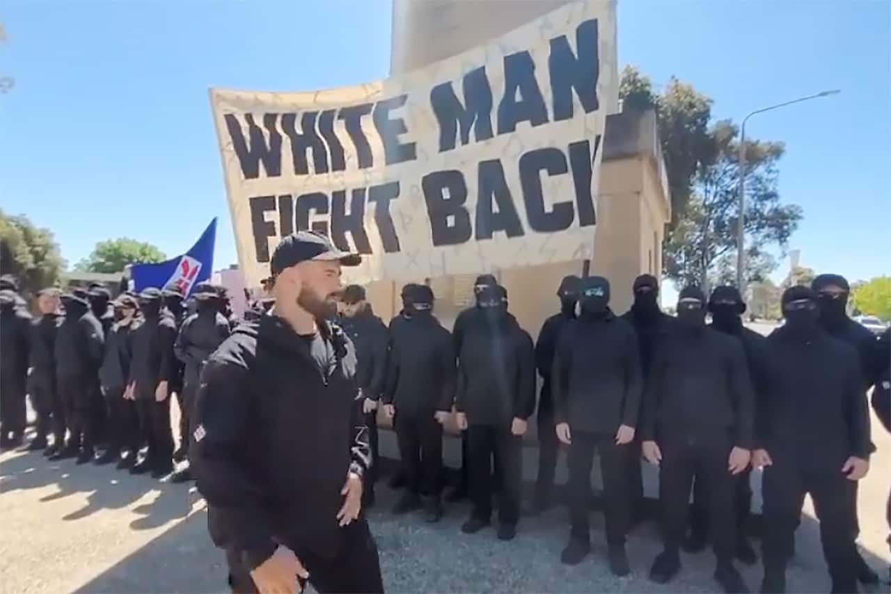 A row of men dressed in all black, most wearing masks. Some are holding a large sign that reads 'white man fight back'