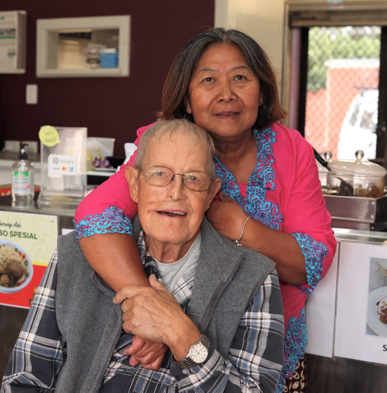 A woman in a pink jacket leans over the shoulder of a seated man in a checked shirt. 