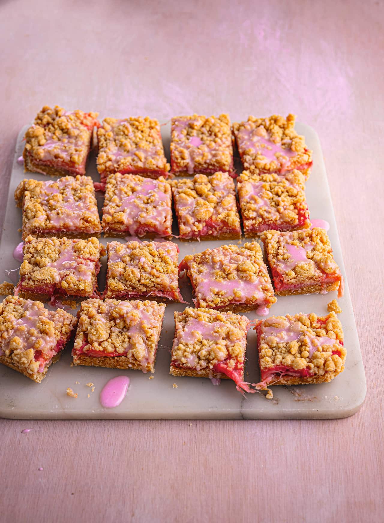 16 squares of a golden-brown slice, studded with rhubarb, sit on a board, drizzled with pink icing. 