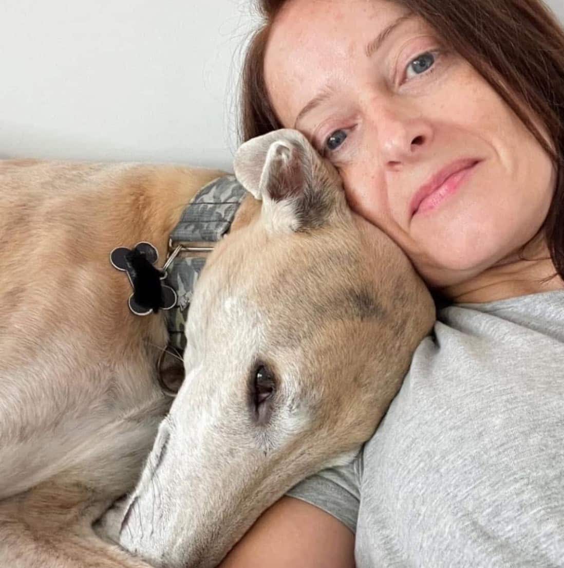 A woman snuggles up with her greyhound. She is smiling slightly. 