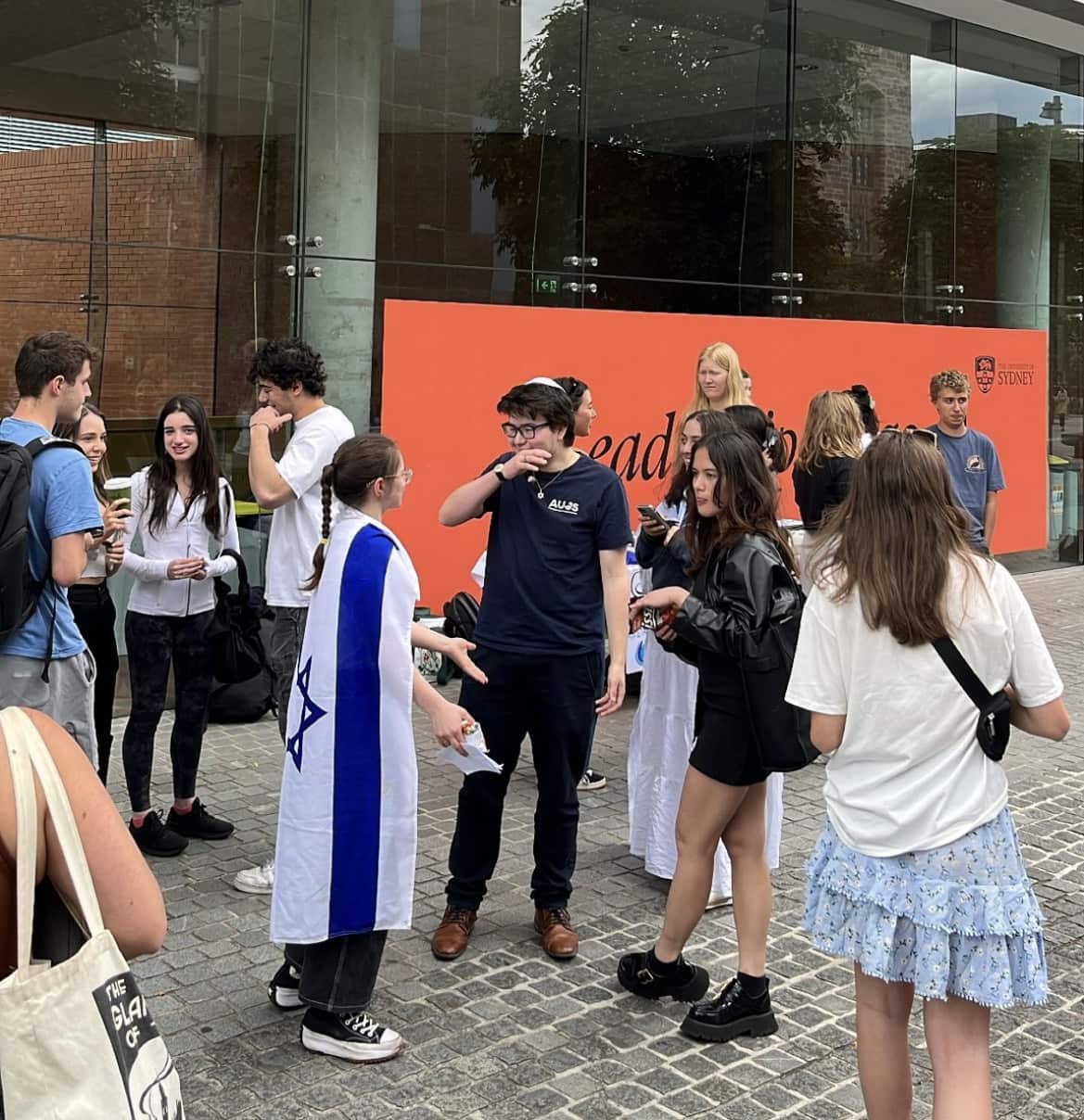 A young woman draped in an Israeli flag speaks to a young man wearing a kippah, amongst a group of other young people.