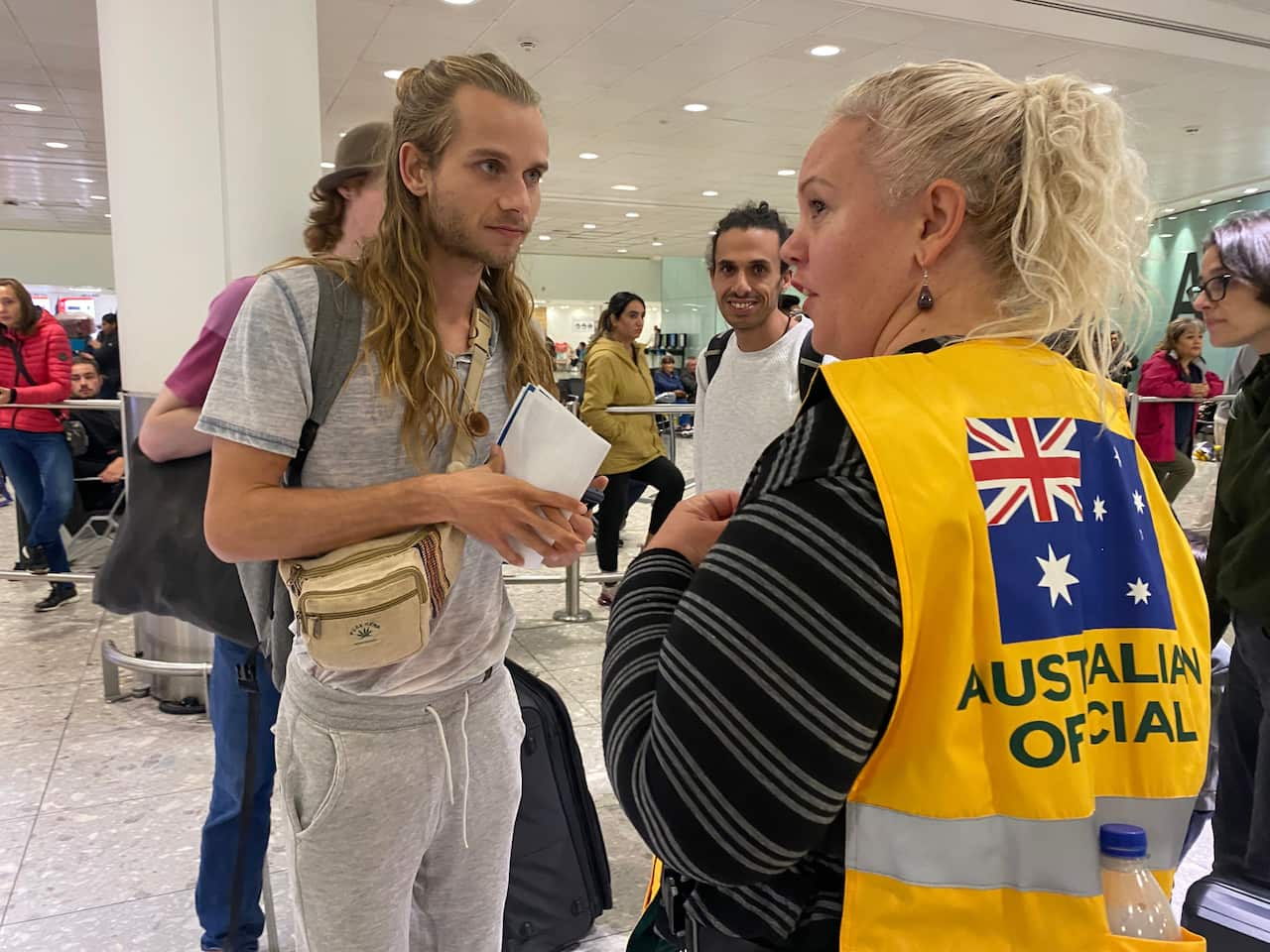 A long-haired man dressed in a grey shirt and pants talks to a woman wearing an "Australian official" vest" 