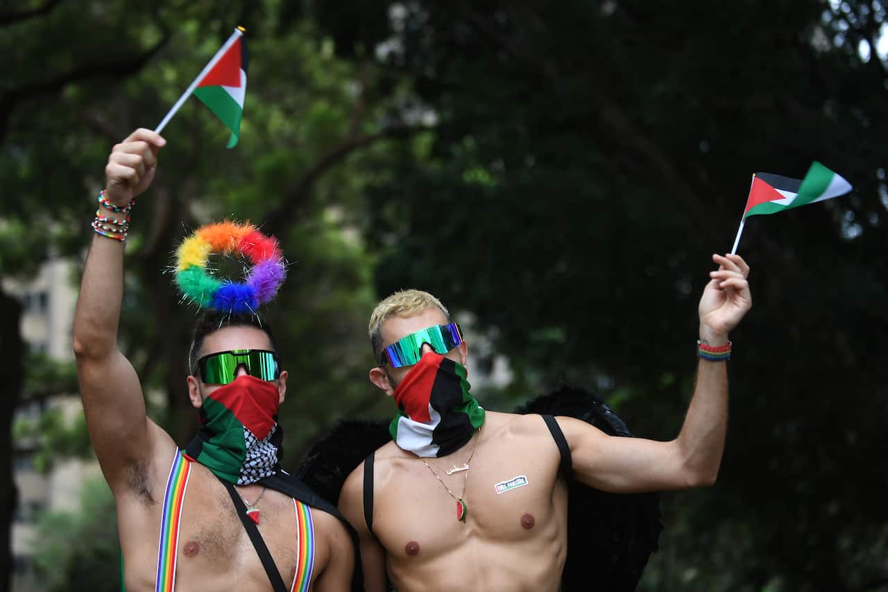 Two men wearing rainbow accessories waving small Palestinian flags