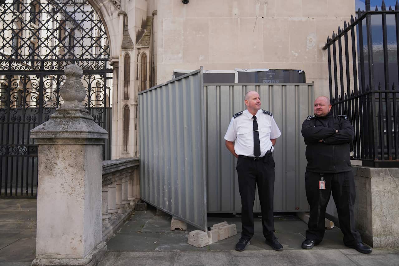 Two security guards standing in front of two metal shed walls which have been placed in front of a stone wall