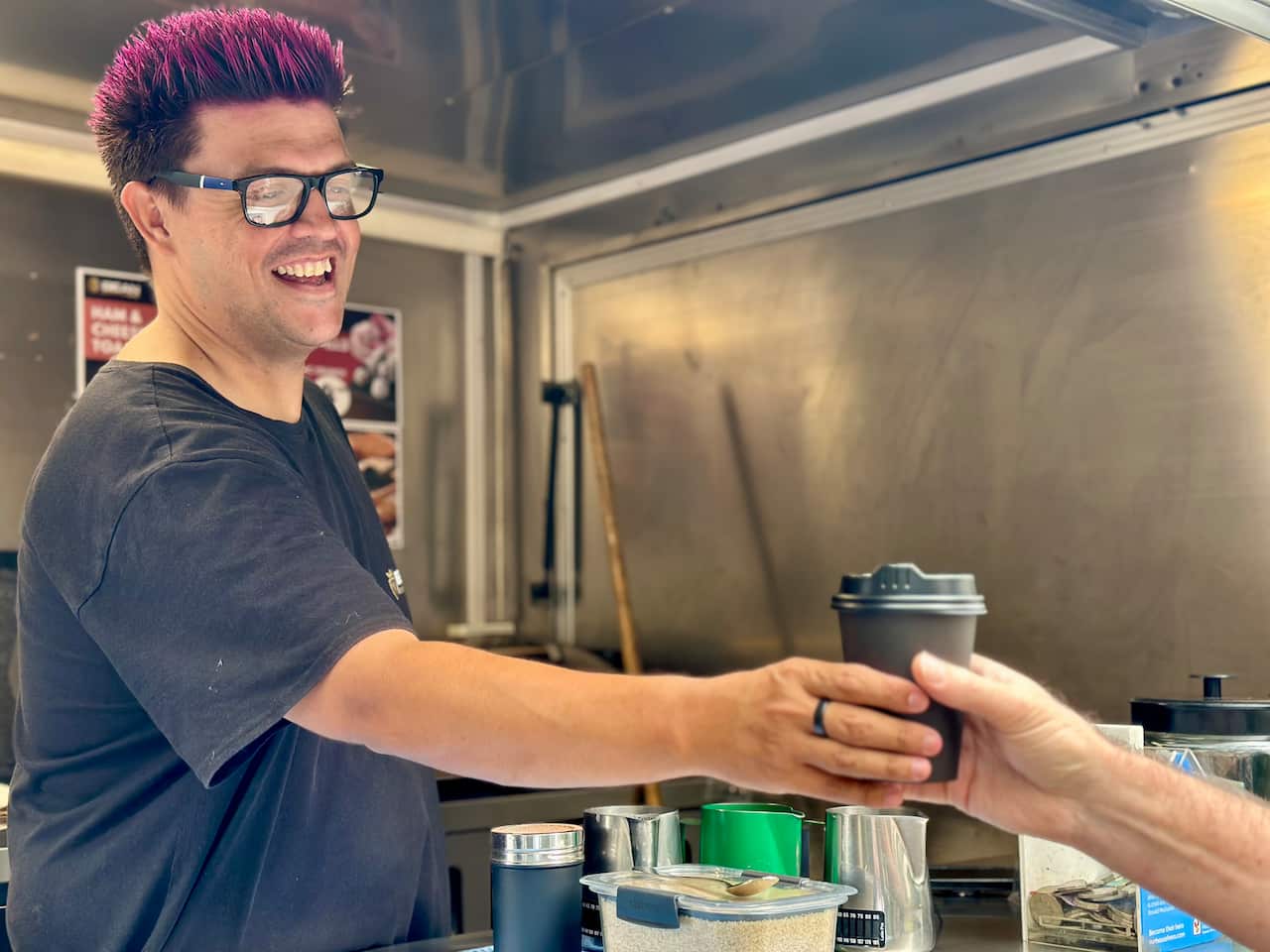 A barista hands a man a coffee while smiling. He is in a silver coffee van