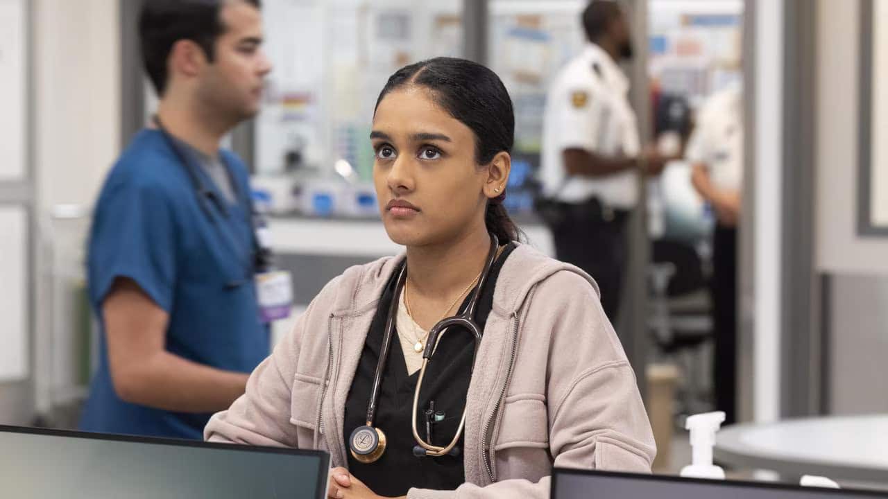 A young woman wearing a stethoscope around her neck. A man in blue scrubs stands behind her in a hospital.