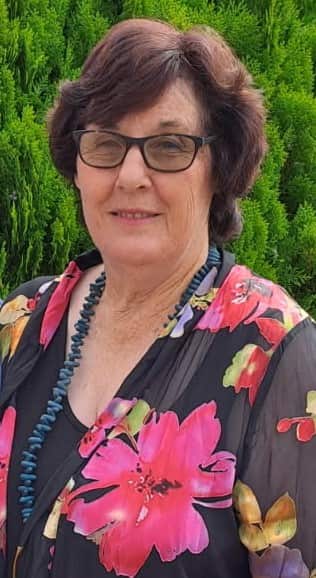 An older woman wearing a floral top, standing in front of hedges.