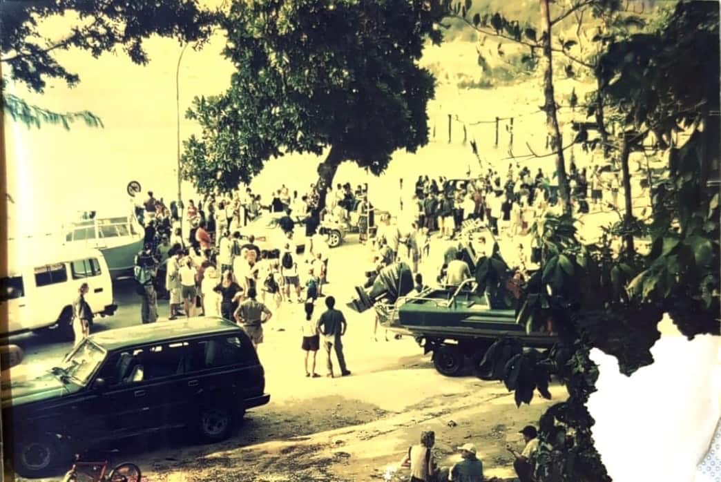 An old grayscale image of a crowd gathered in an open space flanked by trees.