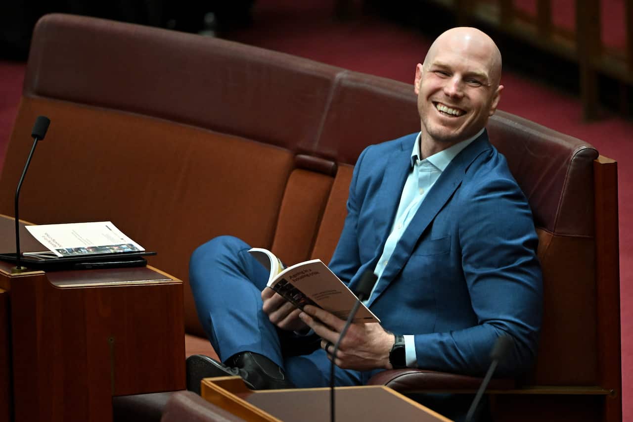 A man in a blue suit with one leg crossed over the other sitting on a red seat