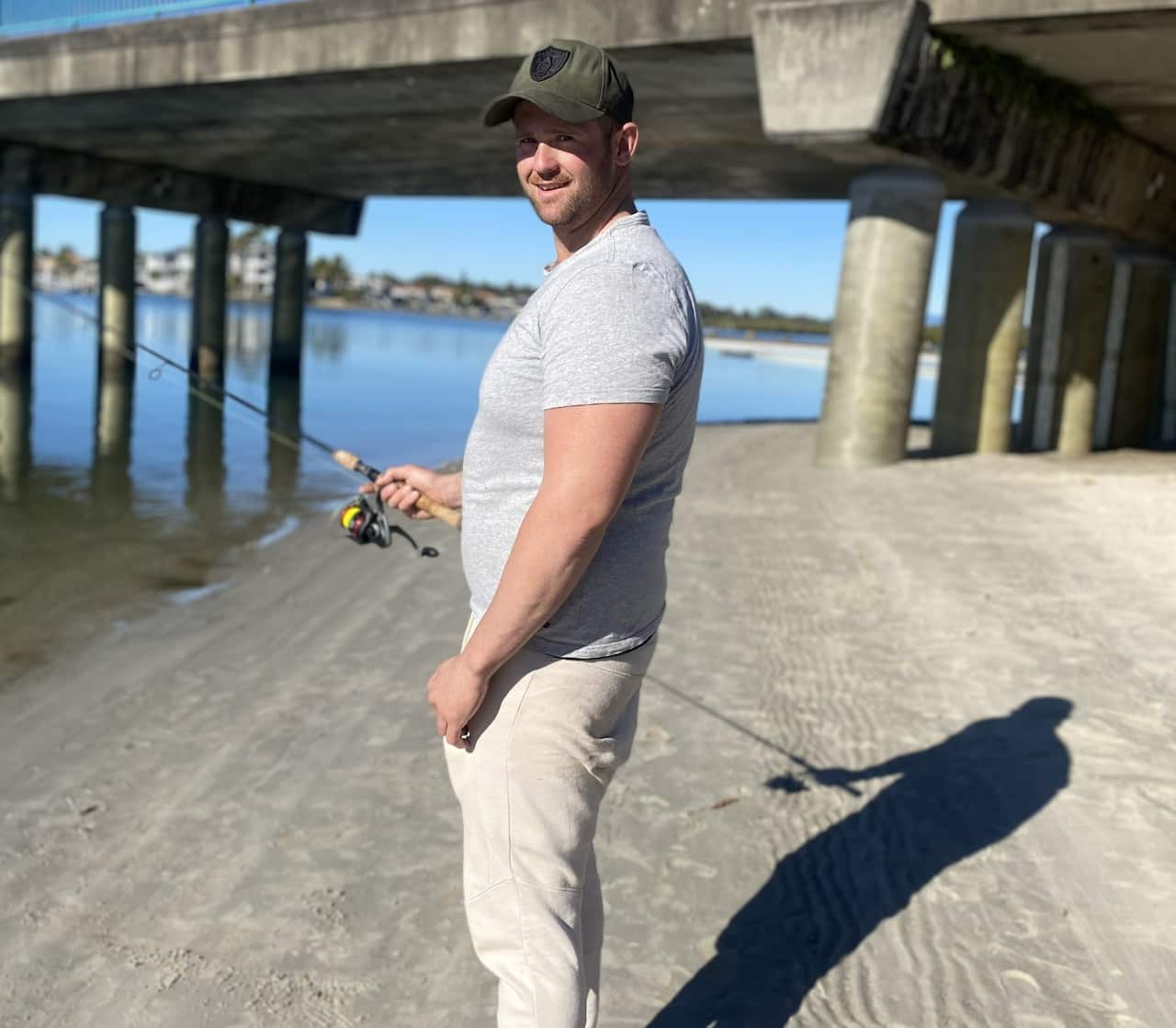 A side profile of a man standing on a beach near a pier holding a fishing rod.