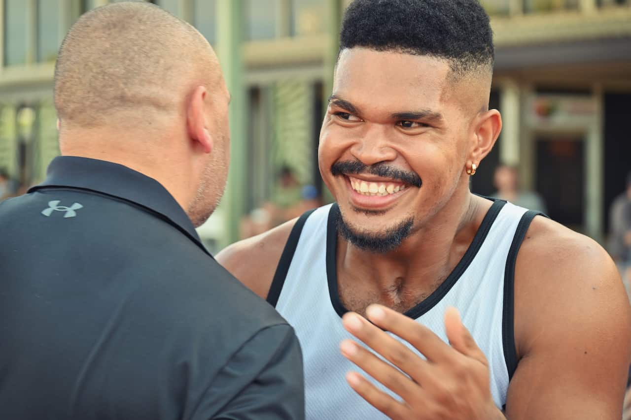 A man in a white singlet smiles broadly as he faces a man in a dark tracksuit top (back to camera).