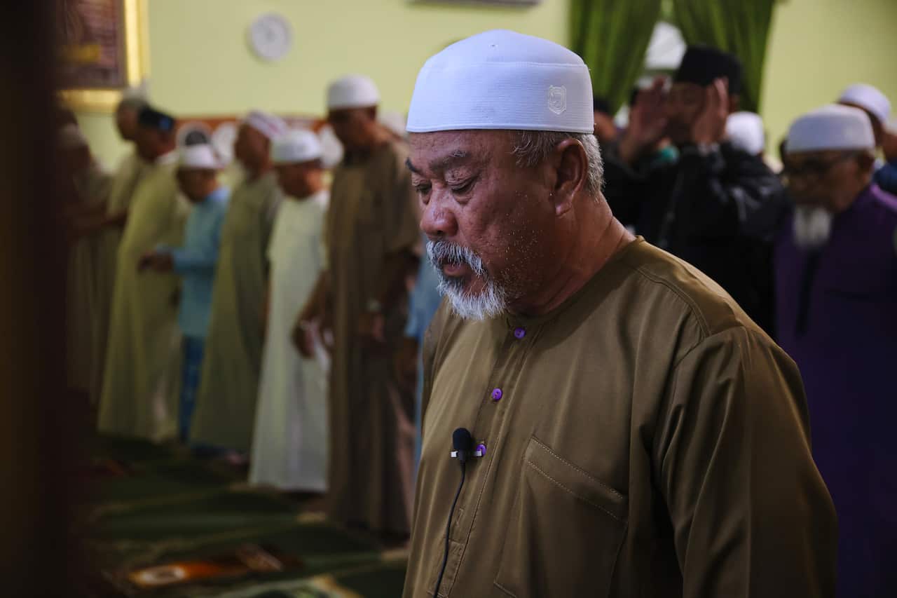 A man wearing a skullcap leads worshippers in prayer inside a mosque, with the congregation standing behind him.