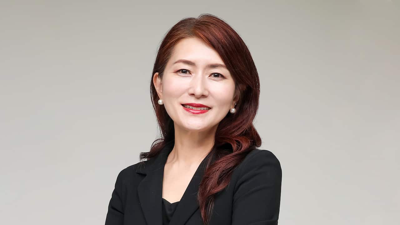 A Korean woman with red dyed hair wearing a black suit and pearl earrings