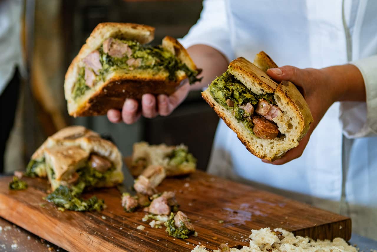 The hands of a person in a white jacket hold two pieces of stuffed bread load above a wooden board, where another piece of stuffed bread sits.  