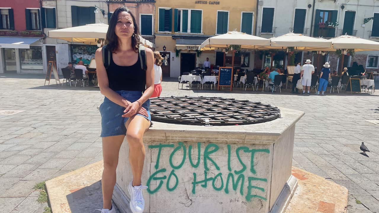 A woman wearing denim shorts and a black tank top posing for a photo sitting on a non-functioning fountain with 'Tourist go home' graffitied on it