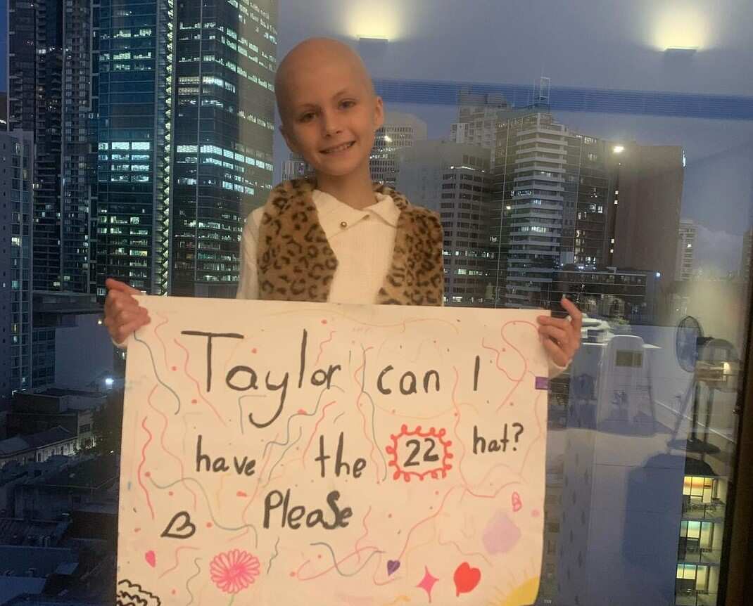 A little girl with no hair is holding a poster that says "Taylor, can I have the 22 hat? Please"