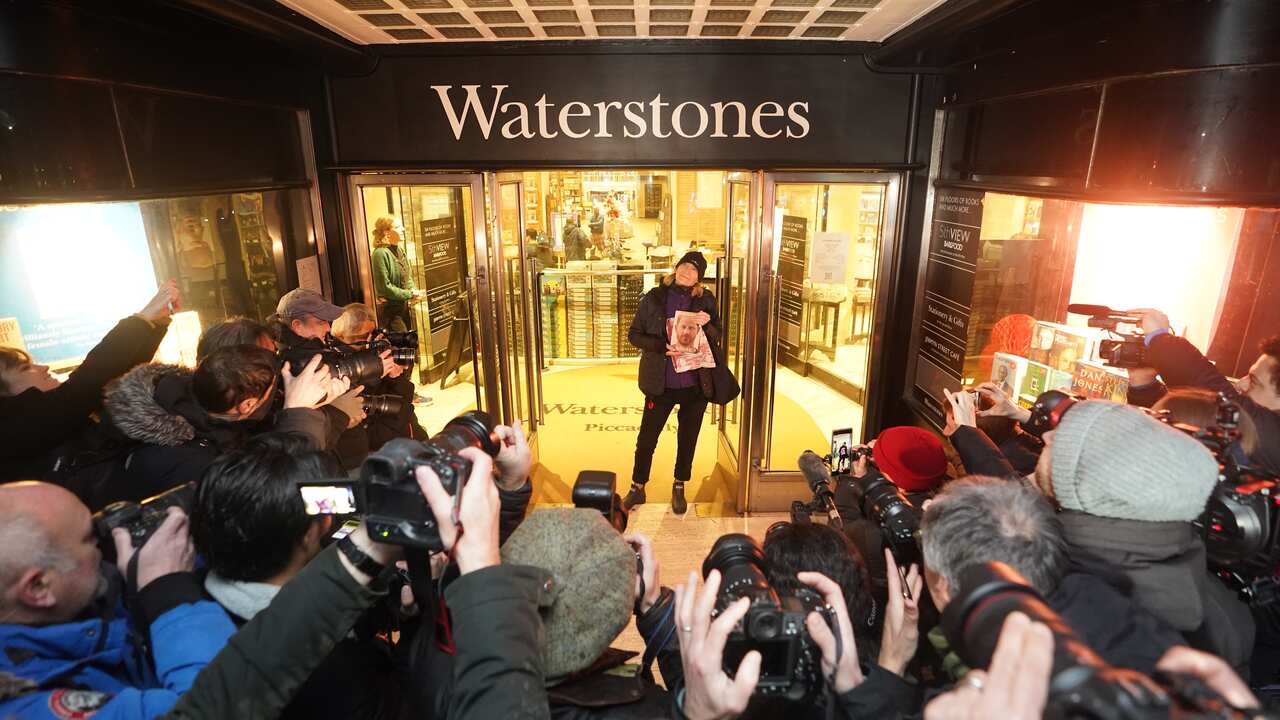 Woman standing in bookshop entry holding book in front of crowd of photographers 