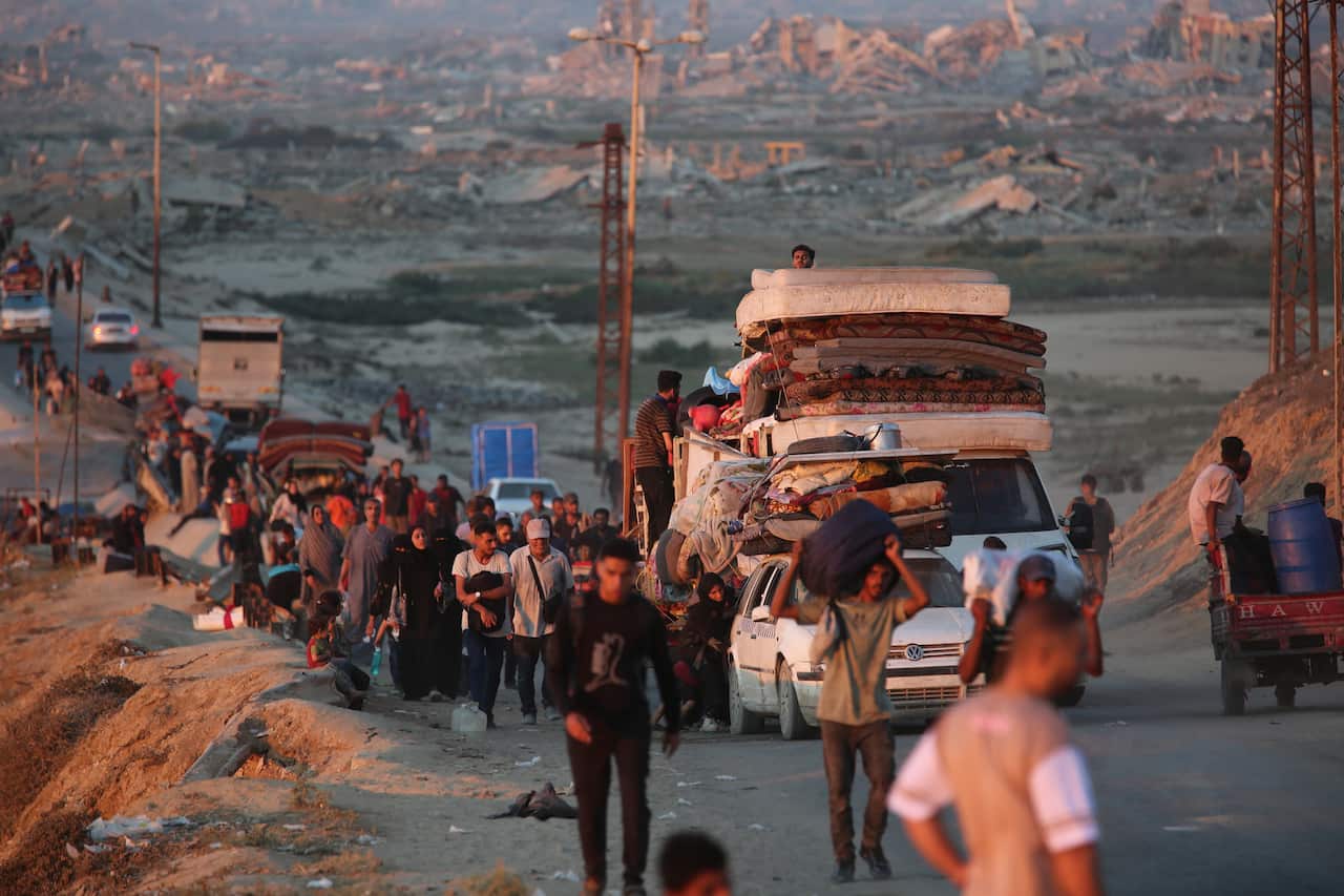 A crowded road shows many people and cars carrying belongings, with mattresses and other items tied to the roofs of vehicles. In the background, a vast landscape of destroyed and flattened buildings stretches to the horizon under a hazy sky.