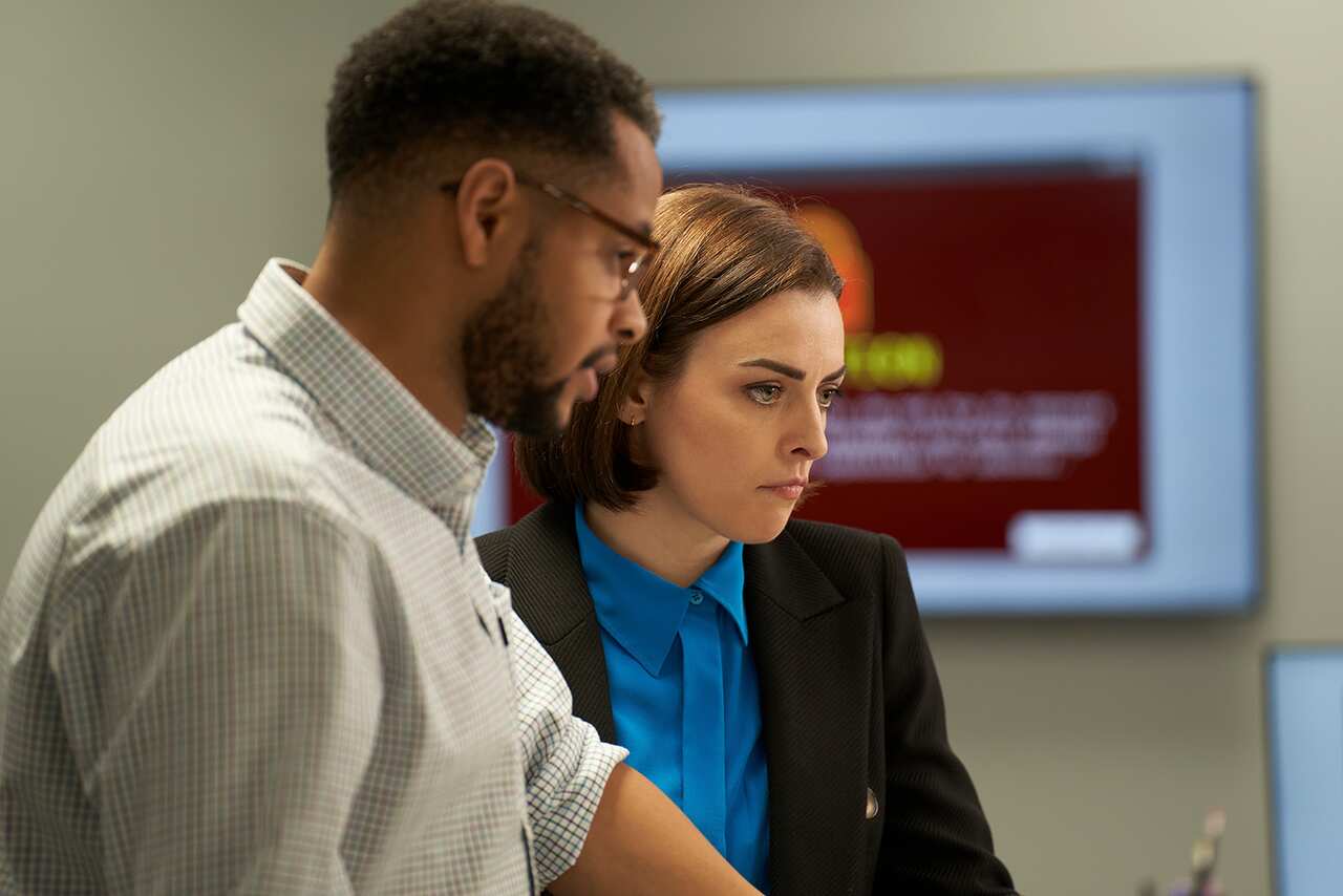 A man in glasses stands side-on. A woman in a bright blue jacket and dark coat stands behind him. They are both looking down and to the side.