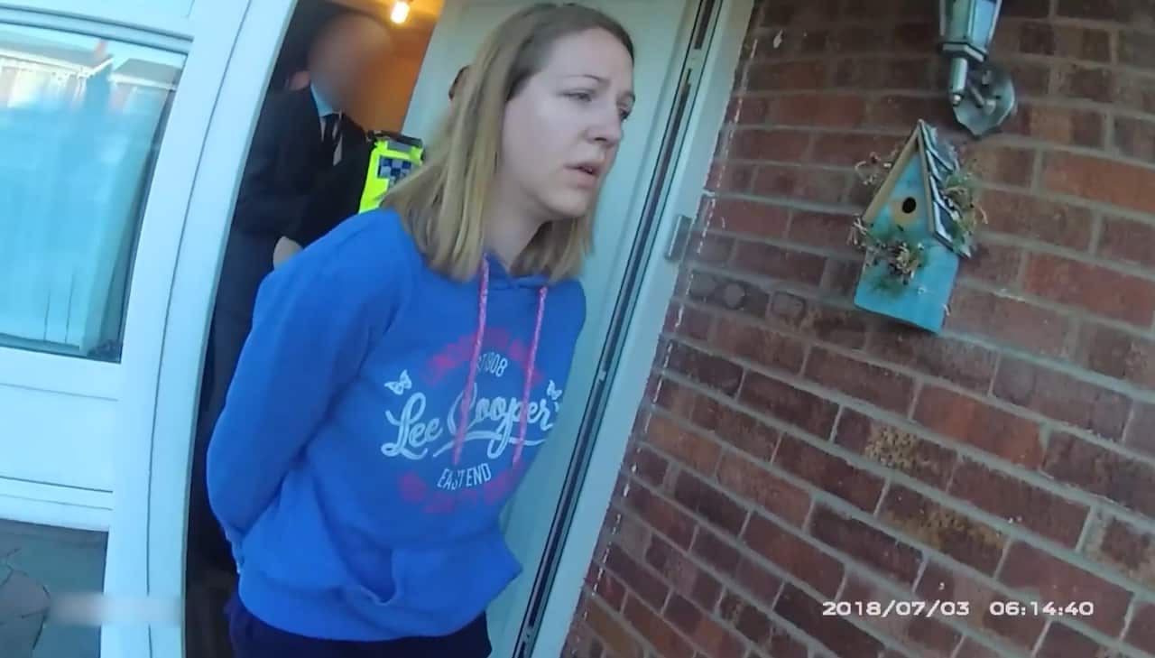 A woman wearing a blue Lee Cooper hoodie and with her hands behind her back is escorted out of a brick house by police officers.