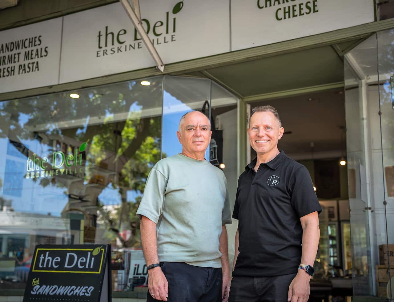 Two men stand next to each other outside a suburban deli.